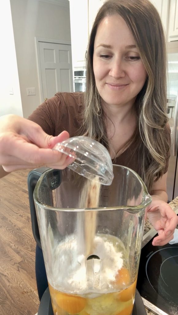 A woman smiles while pouring sugar from a small bowl into a blender filled with eggs and flour in a kitchen.