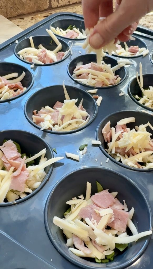 A hand sprinkles shredded cheese over chopped ham and spinach in a muffin tin, preparing individual portions for baking.