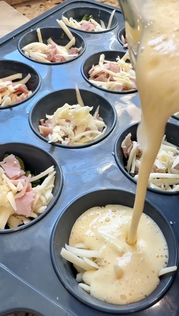 A muffin tin filled with shredded cheese and ham pieces is being filled with beaten eggs, preparing for baking, likely to make mini egg muffins or quiches.