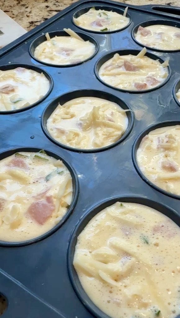 A close-up of a muffin tin filled with an uncooked egg mixture containing chopped ham, cheese, and vegetables, ready to be baked.