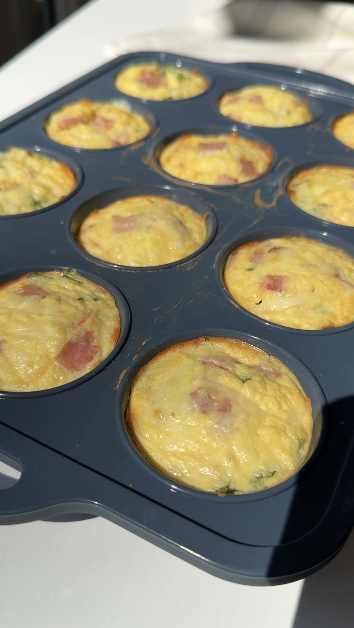 A close-up of a muffin tin filled with baked egg bites containing visible pieces of ham and herbs, sitting on a surface in bright sunlight.