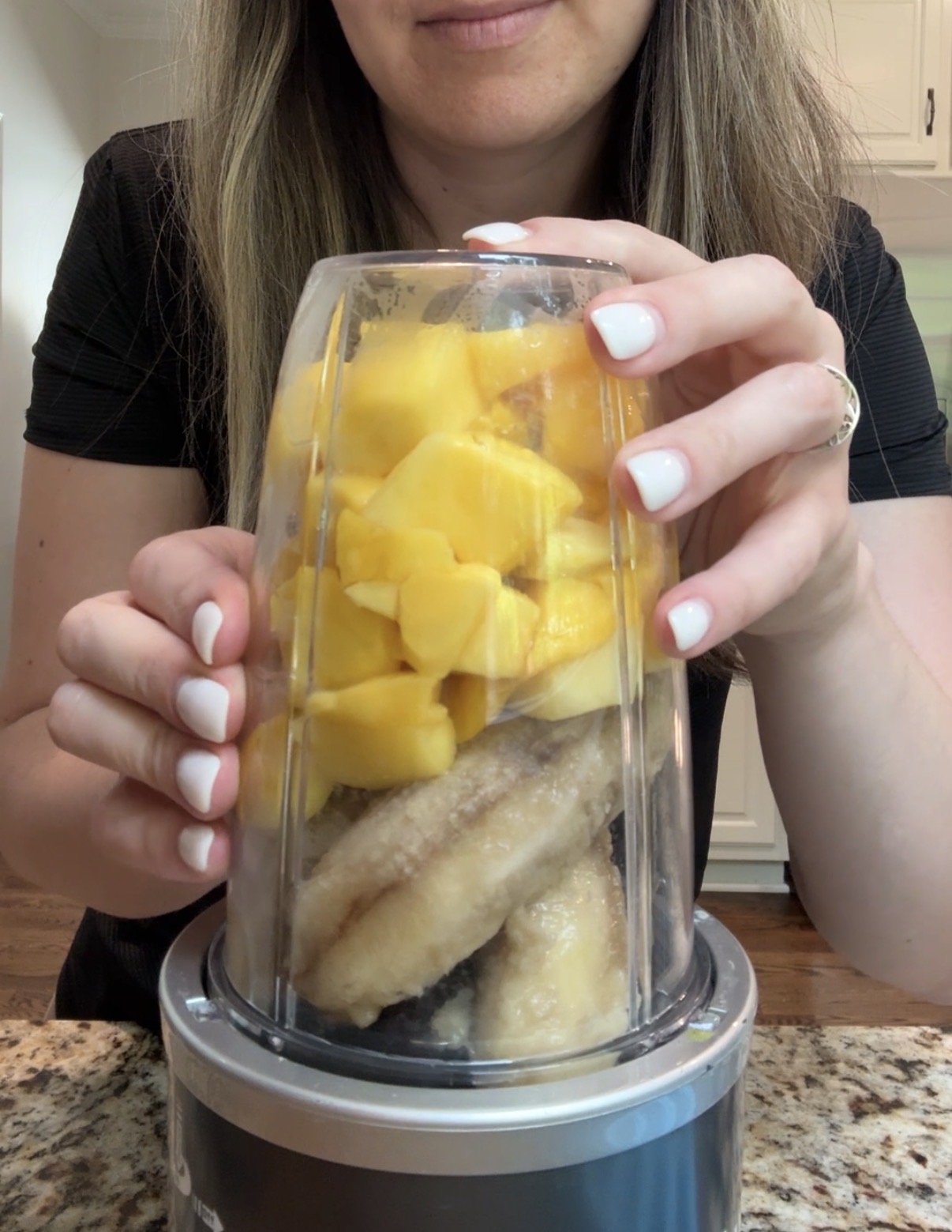A woman holds a blender cup filled with frozen bananas and chopped mango, preparing to blend them on a kitchen counter.