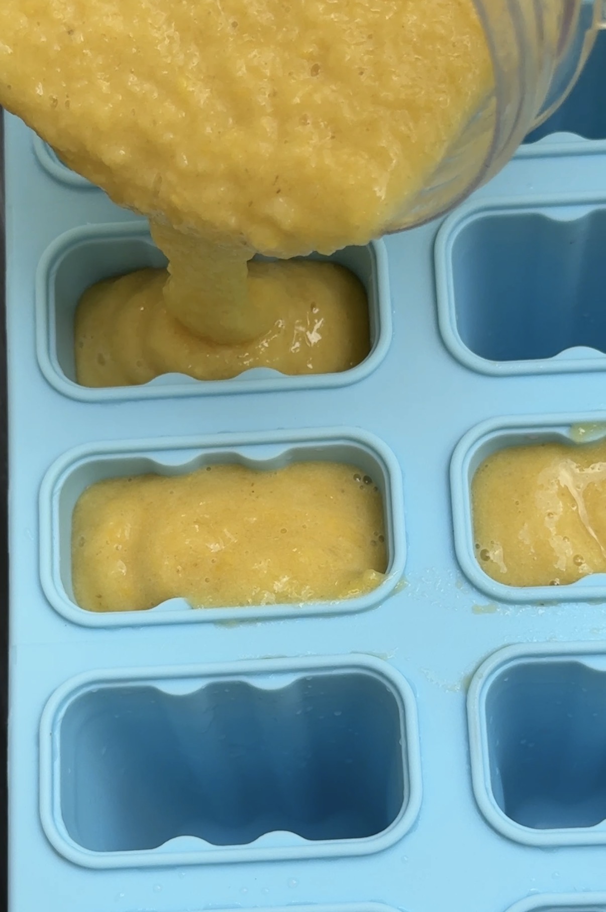 Yellow mixture being poured from a container into a blue silicone ice cube tray with rectangular compartments.
