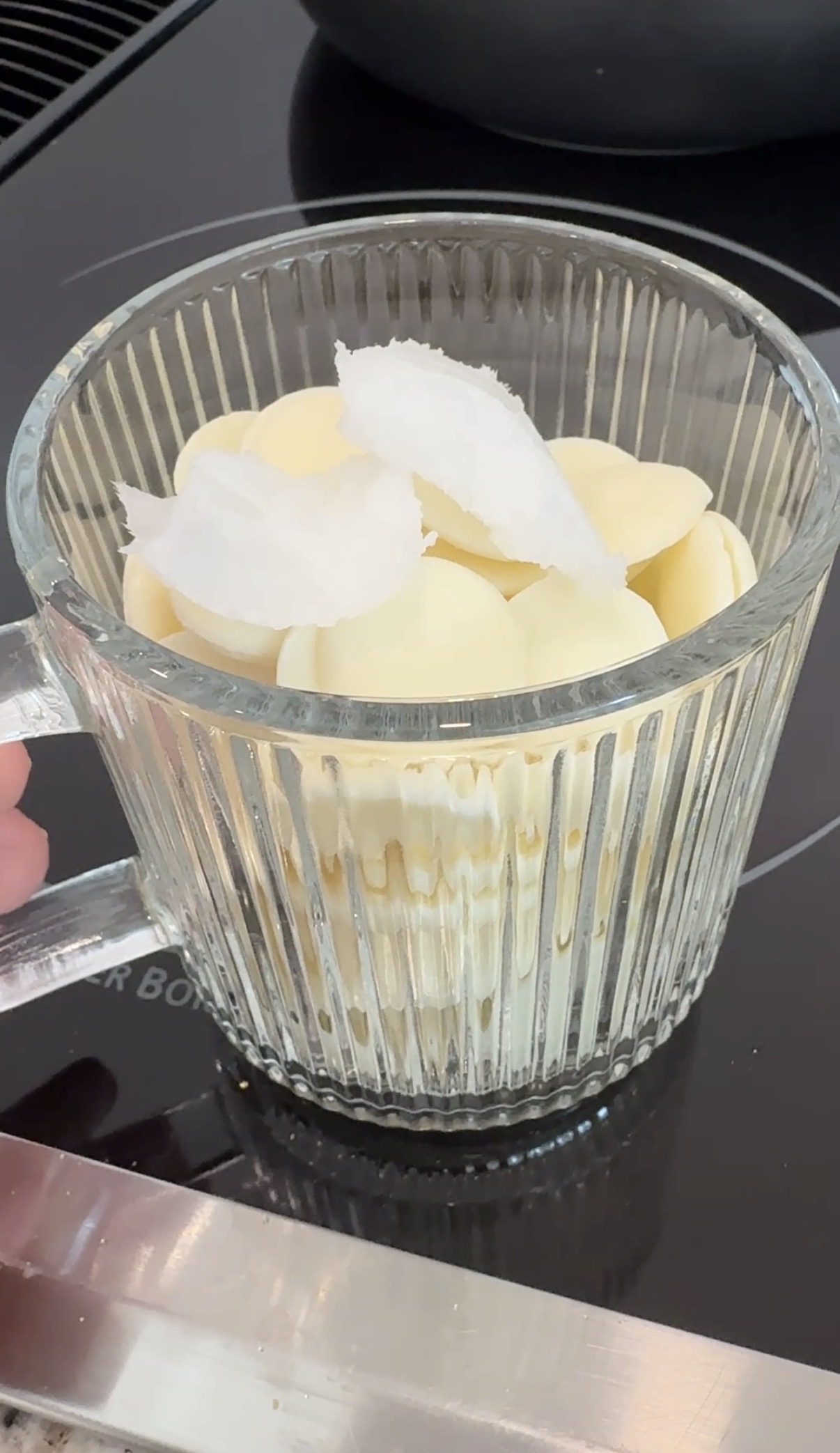 A clear glass mug filled with white chocolate melting wafers and a few chunks of solid coconut oil sits on a stovetop.