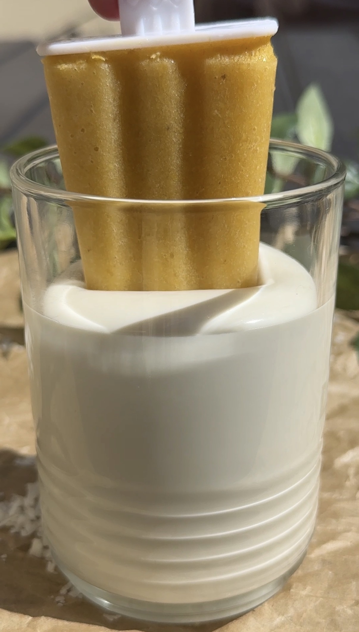 A yellow popsicle is being dipped into a glass filled with creamy white liquid. The background is softly blurred with hints of greenery.