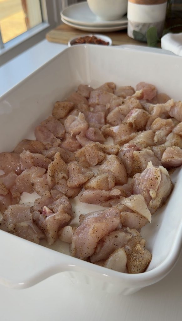 Cubed raw chicken pieces, seasoned with spices, are spread evenly in a white rectangular baking dish, ready to be cooked. A window and countertop items are blurred in the background.