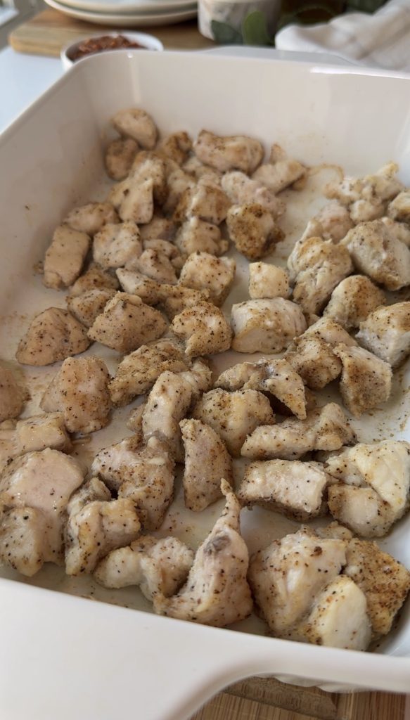 Seasoned, cooked chunks of chicken breast are spread out in a white baking dish. The chicken pieces appear lightly browned and evenly coated with spices.