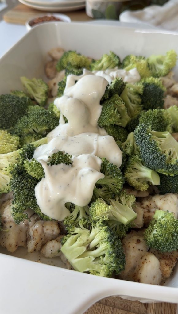 A close-up of a casserole dish filled with seasoned chunks of chicken, fresh broccoli florets, and a generous layer of creamy white sauce on top.