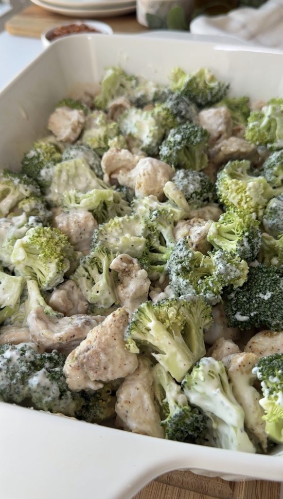 A close-up of a dish filled with broccoli florets and pieces of chicken, all coated in a creamy white sauce and seasoned with black pepper.