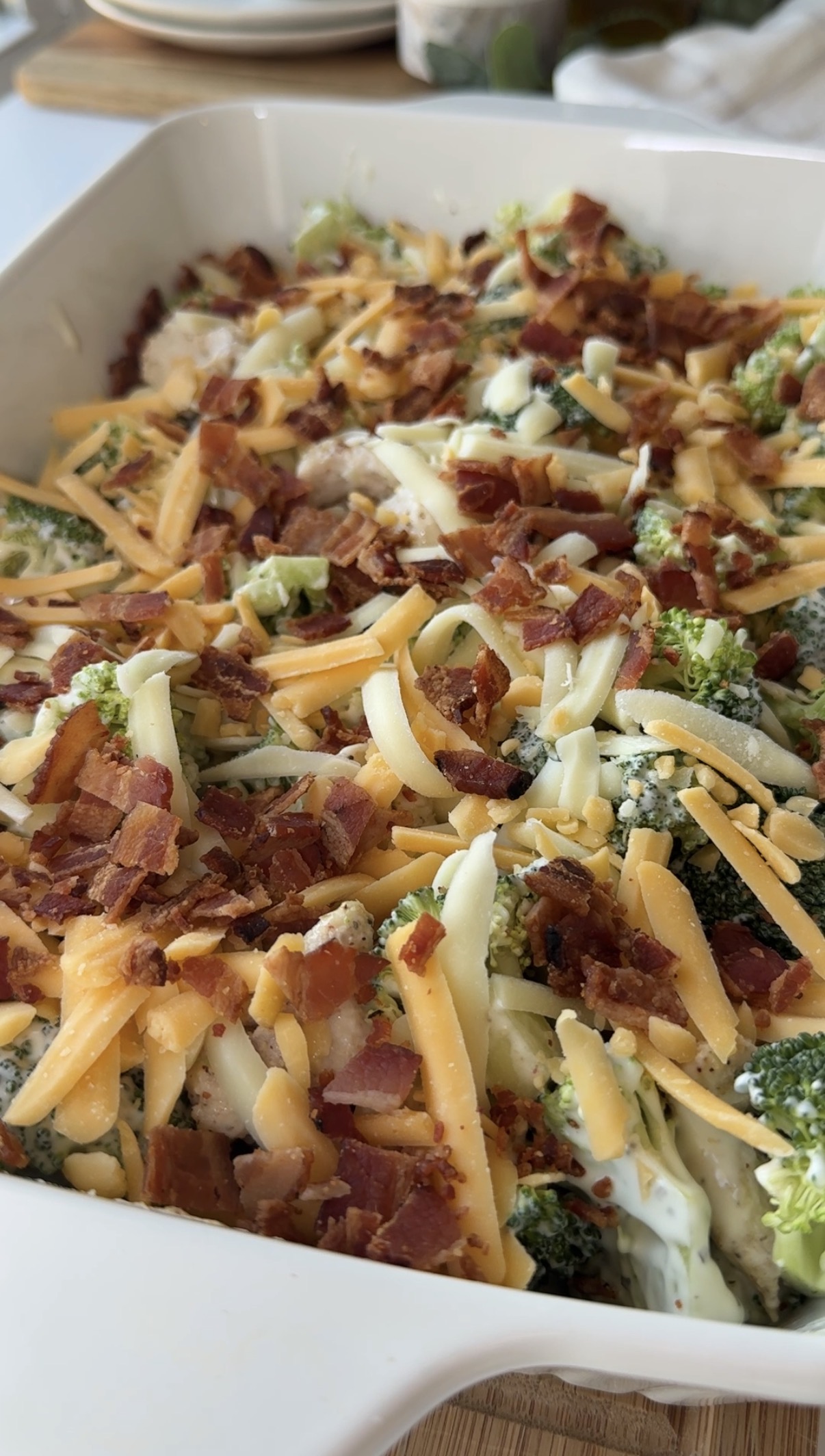 A close-up of a casserole dish filled with broccoli, shredded cheddar cheese, bacon bits, and a creamy sauce, ready to be baked.