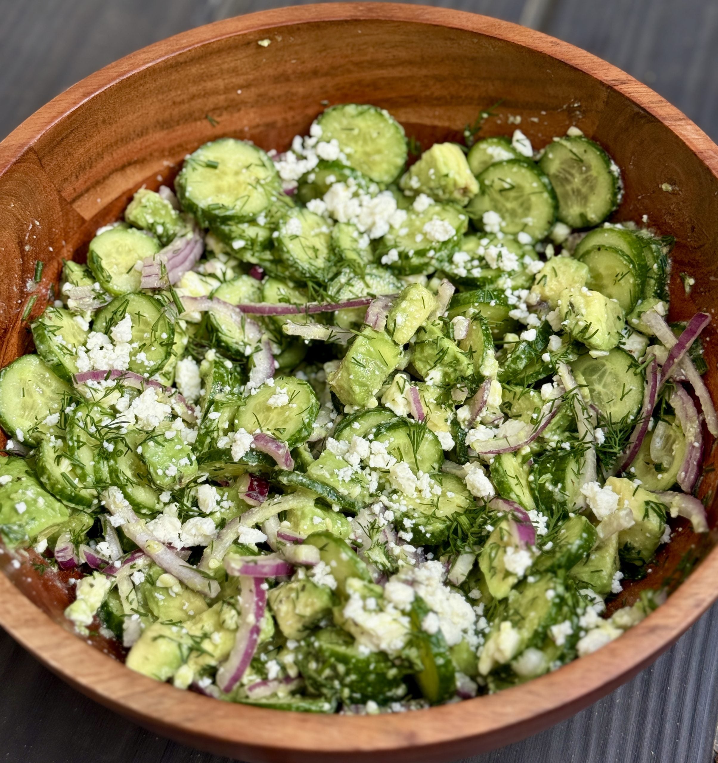A wooden bowl filled with cucumber salad, featuring sliced cucumbers, red onion, fresh dill, crumbled feta cheese, and avocado chunks, all tossed together on a dark wooden surface.