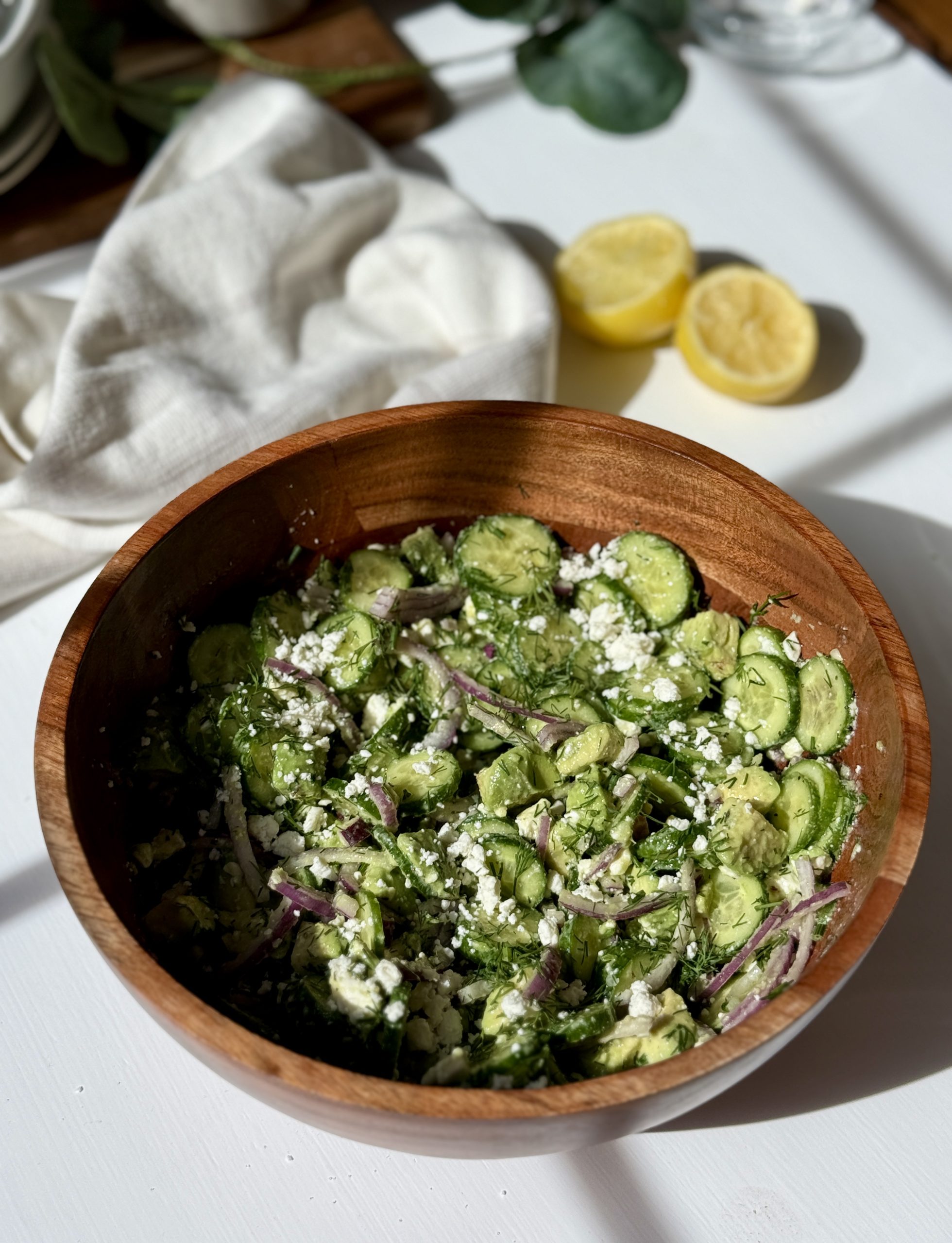 A wooden bowl filled with a fresh salad of sliced cucumbers, red onions, crumbled feta cheese, and herbs sits on a white surface. A white cloth and two lemon halves are in the background.