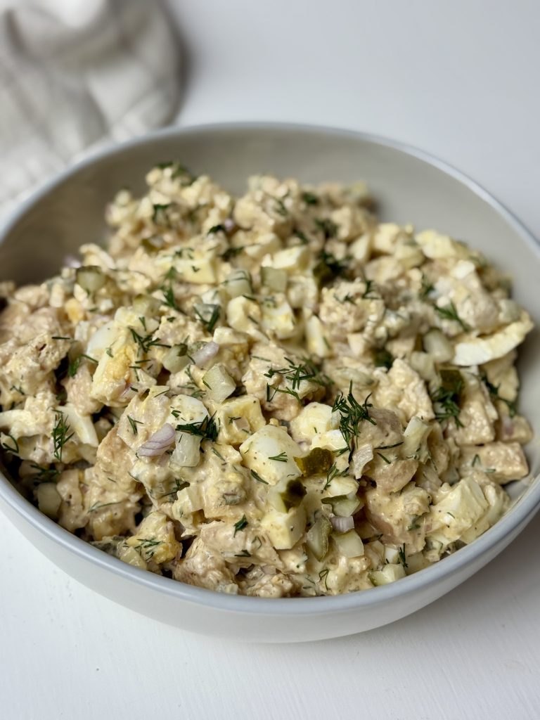 A bowl filled with a chicken salad mixed with eggs, dill, pickles, shallots, yogurt, mayo, and dijon mustard.