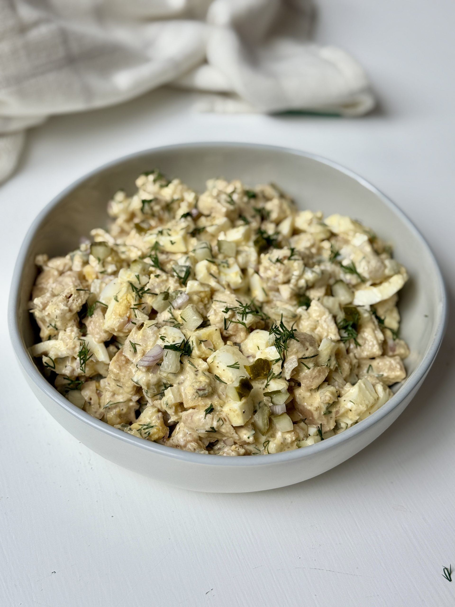 A bowl filled with a chicken salad mixed with eggs, dill, pickles, shallots, yogurt, mayo, and dijon mustard.