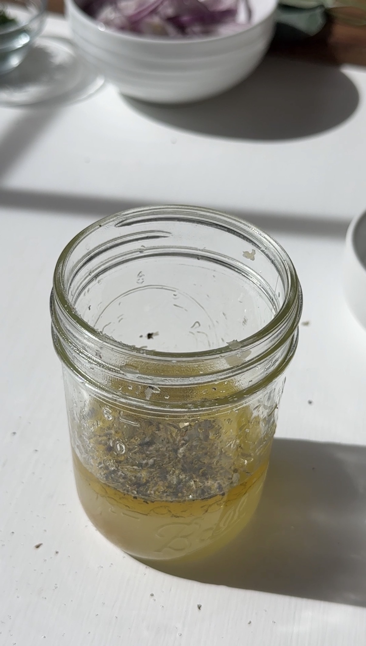 A glass jar containing a yellow liquid with herbs or seasoning floating on top sits on a white surface, with bowls and kitchen items blurred in the background.
