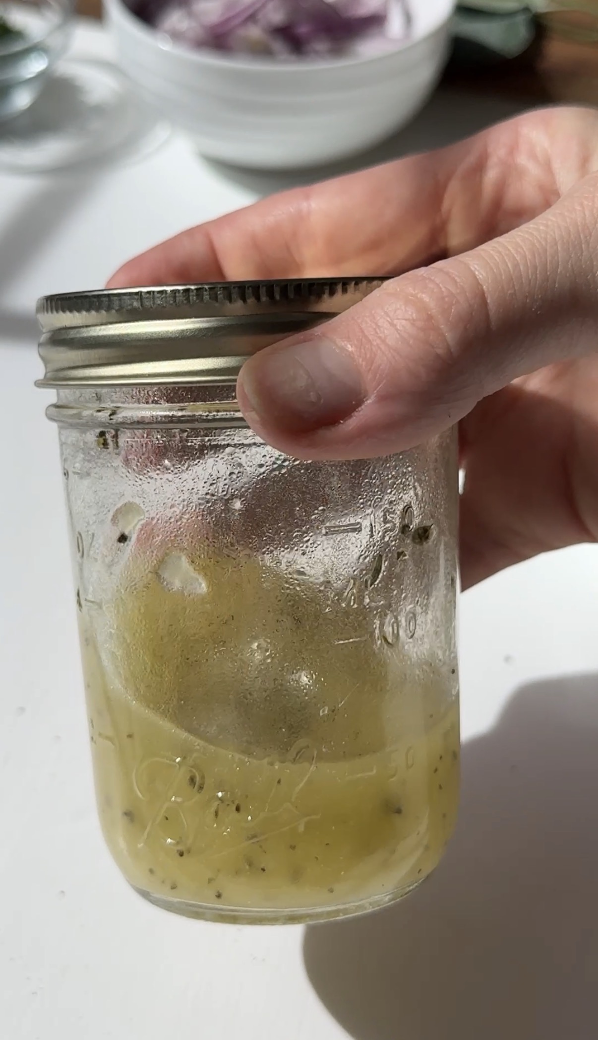 A glass jar containing a yellow liquid with herbs or seasoning floating on top sits on a white surface, with bowls and kitchen items blurred in the background.
