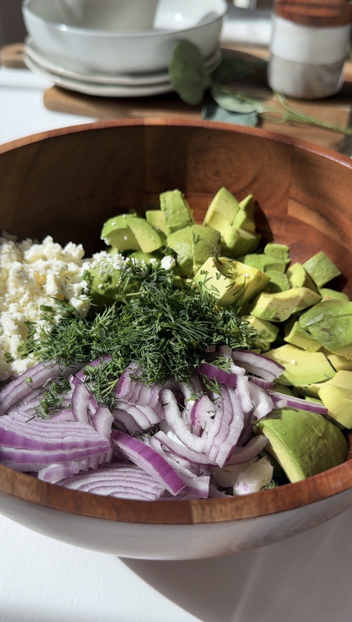 A wooden bowl filled with sliced red onions, cubed avocado, crumbled cheese, and fresh chopped herbs, sitting on a sunlit table.