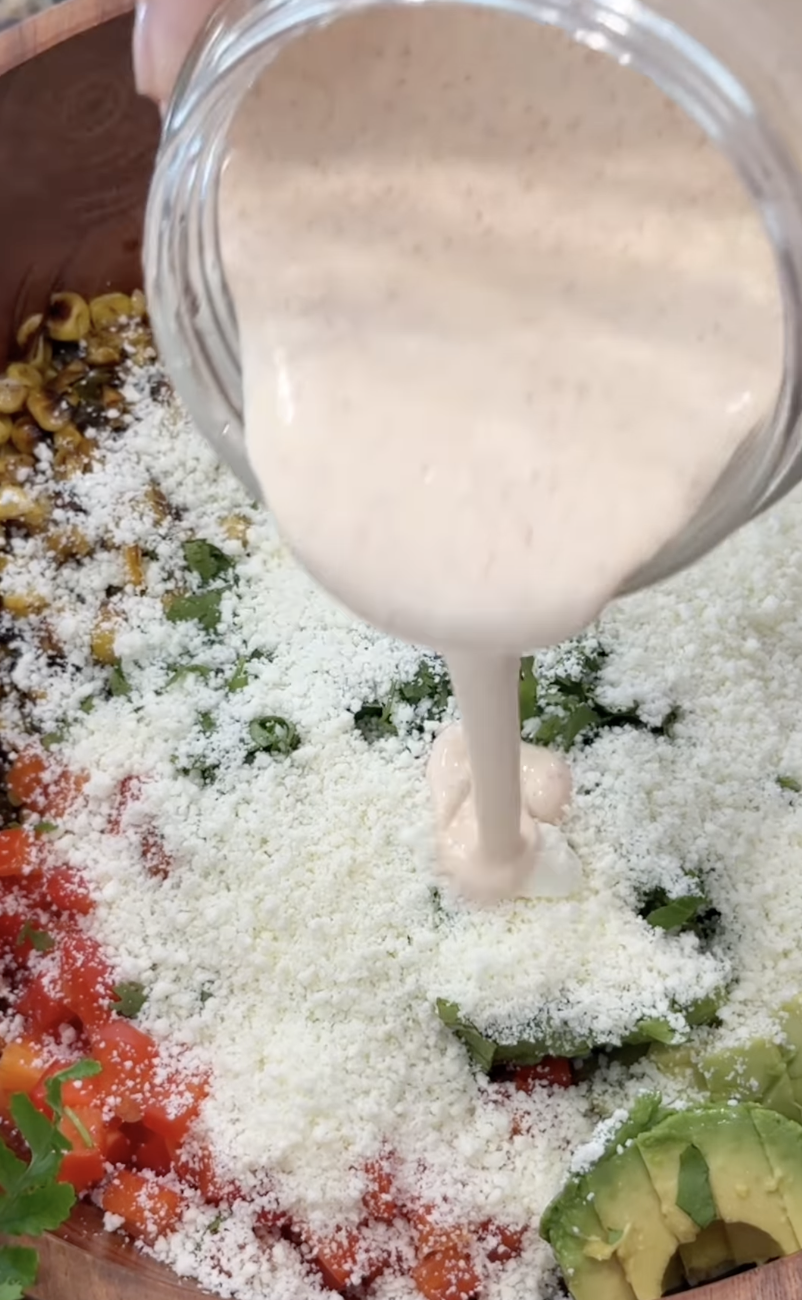 A creamy dressing is being poured from a jar onto a salad topped with grated cheese, diced tomatoes, corn, greens, and avocado slices.