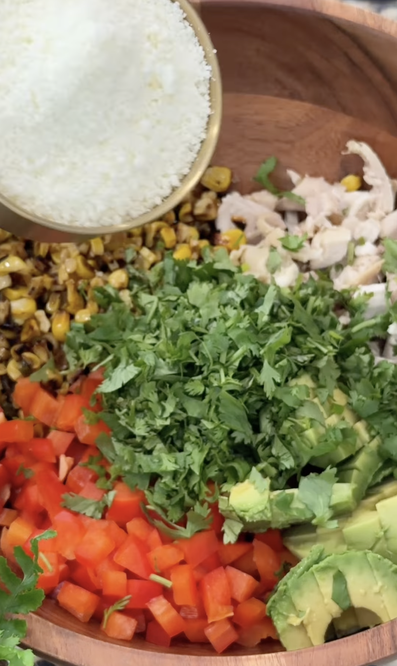 A wooden bowl filled with chopped red bell pepper, avocado slices, cilantro, chicken, roasted corn, and a small bowl of grated cheese being added on top.