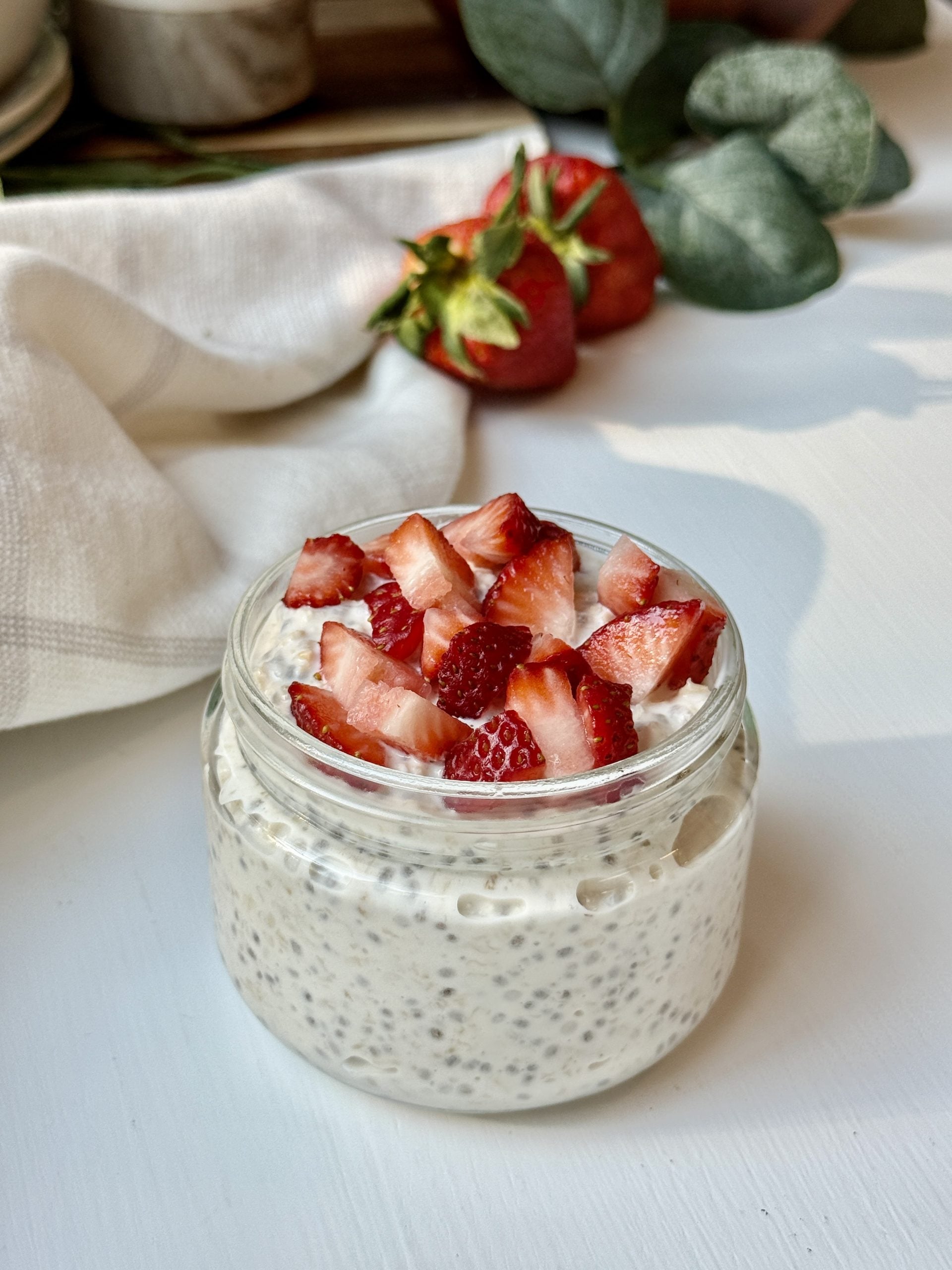 A glass jar filled with chia seed pudding, topped with chopped fresh strawberries, sits on a white surface. In the background, a white cloth and whole strawberries are visible.