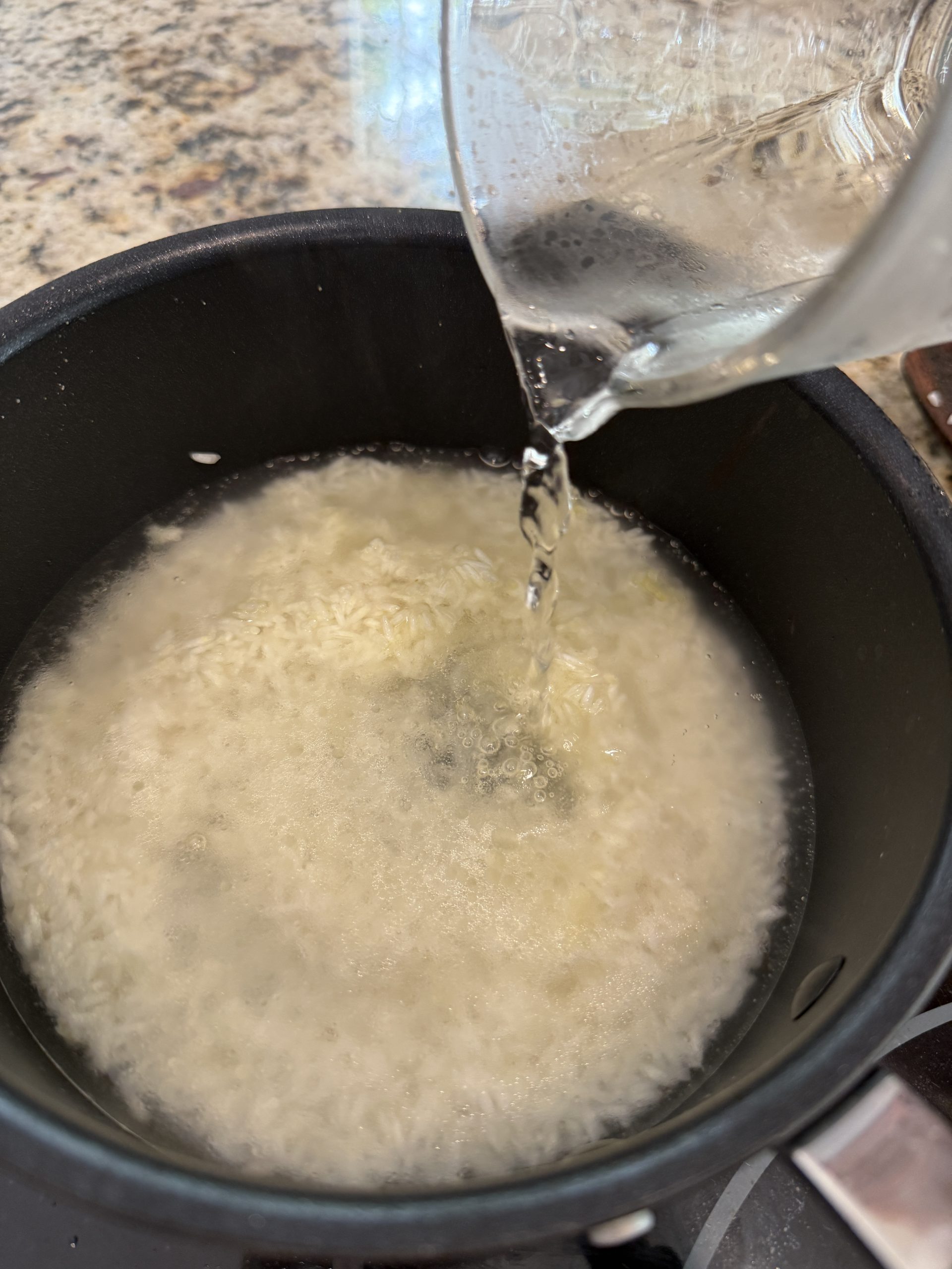 A close-up of water being poured from a glass measuring cup into a black pot of uncooked rice on a stovetop, with the rice beginning to soak.