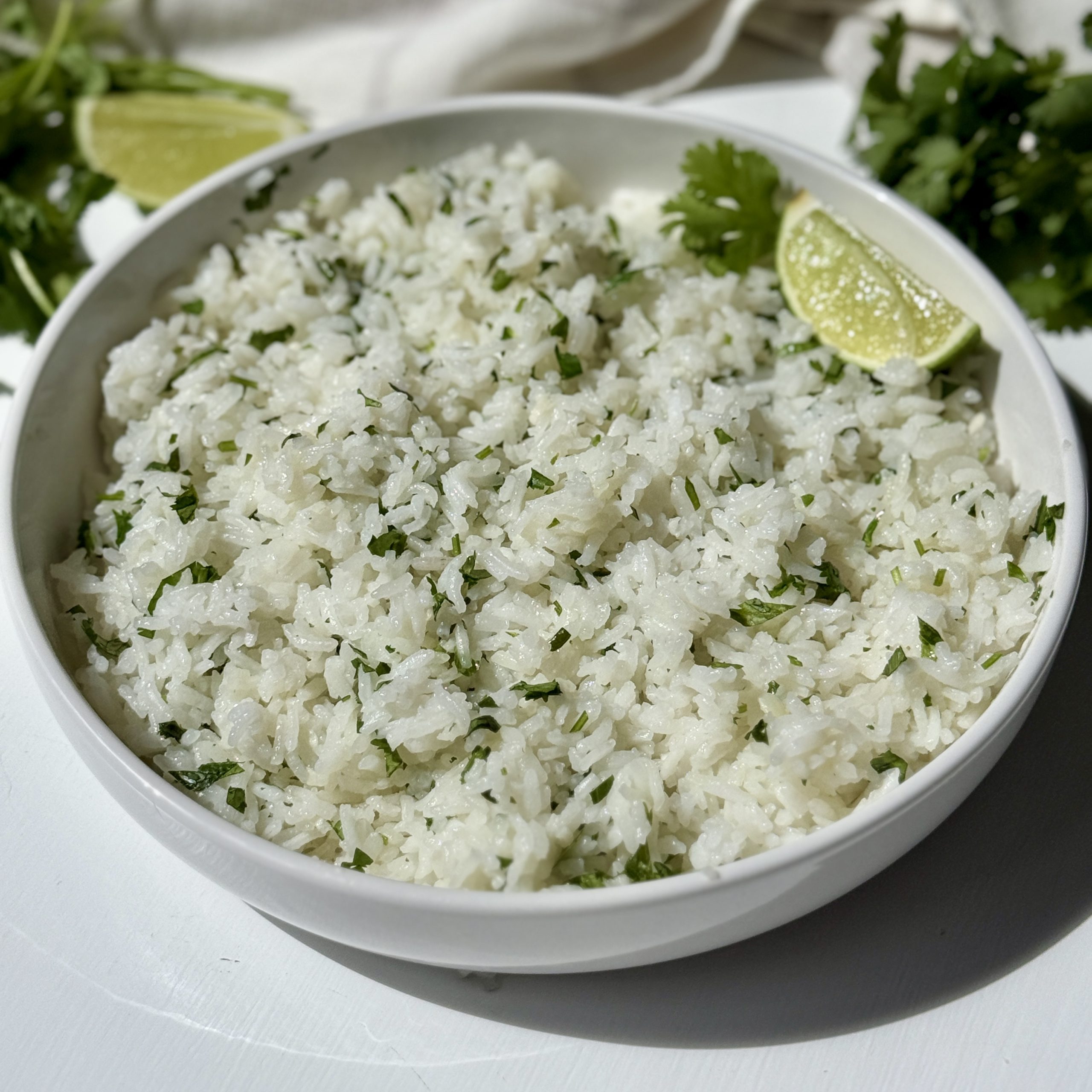 A white bowl filled with fluffy white rice mixed with chopped cilantro, garnished with a lime wedge, sits on a white surface with fresh cilantro and a lime in the background.