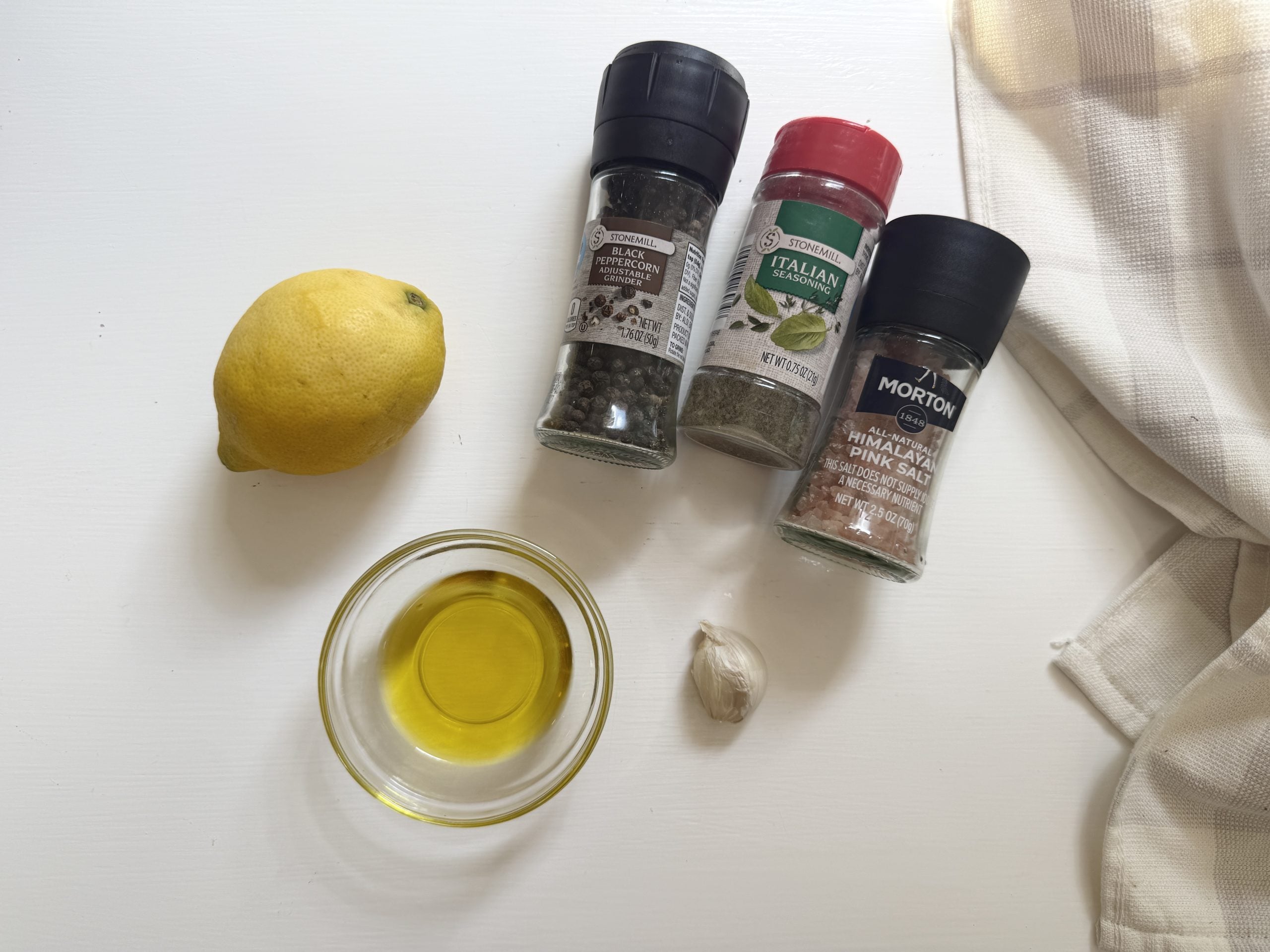 A lemon, a glass bowl of olive oil, a garlic clove, black pepper, Italian seasoning, pink salt, and a white-striped cloth on a white surface.