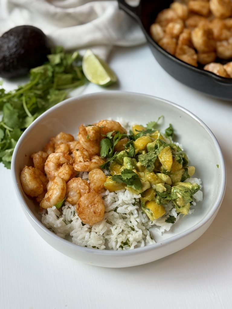 A white bowl filled with cilantro rice, seasoned shrimp, and a mango avocado salsa, with fresh cilantro, a lime wedge, and an avocado in the background. A skillet with cooked shrimp is also partially visible.
