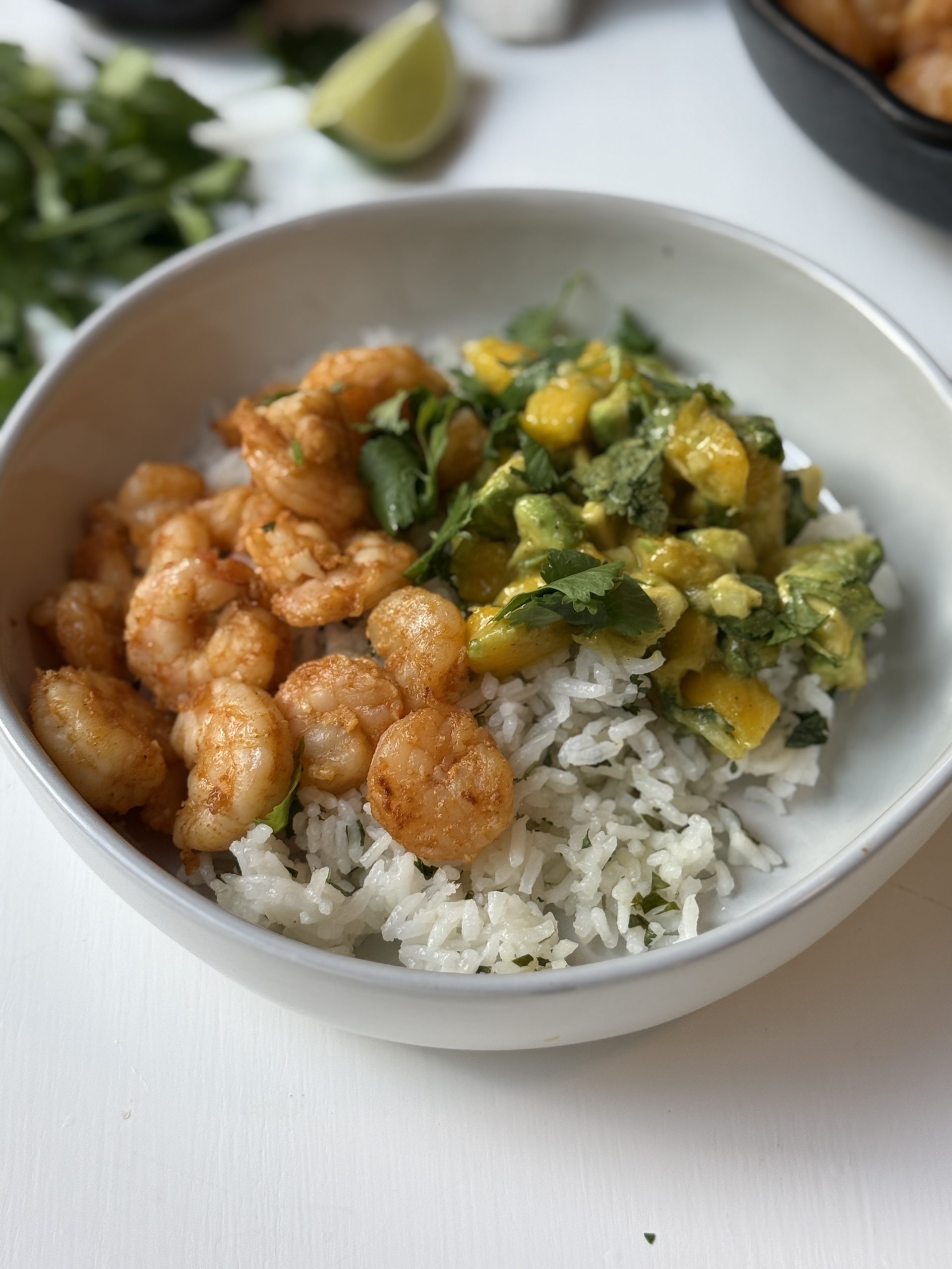 A white bowl filled with cooked shrimp, cilantro rice, and a mango-avocado salsa, garnished with fresh cilantro, set on a white surface.