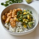 A white bowl filled with cooked shrimp, cilantro rice, and a mango-avocado salsa, garnished with fresh cilantro, set on a white surface.