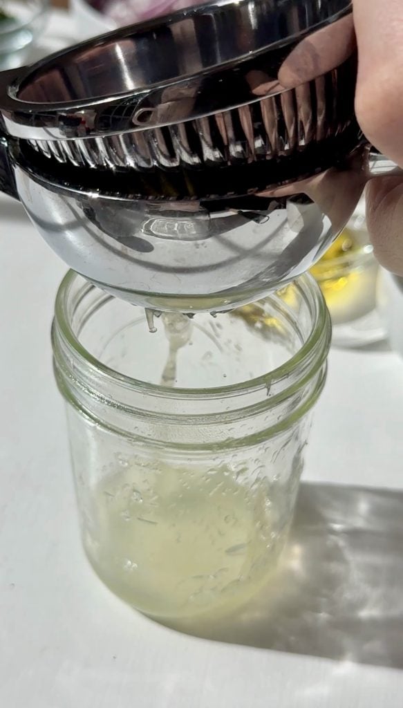 A metal citrus squeezer is being used to juice a lemon or lime over a glass jar. Fresh juice is dripping into the jar, and sunlight casts shadows on the white surface below.