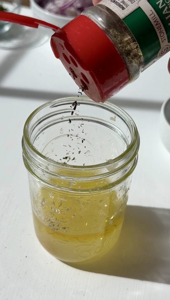 A hand sprinkles dried Italian seasoning from a spice container into a glass jar.