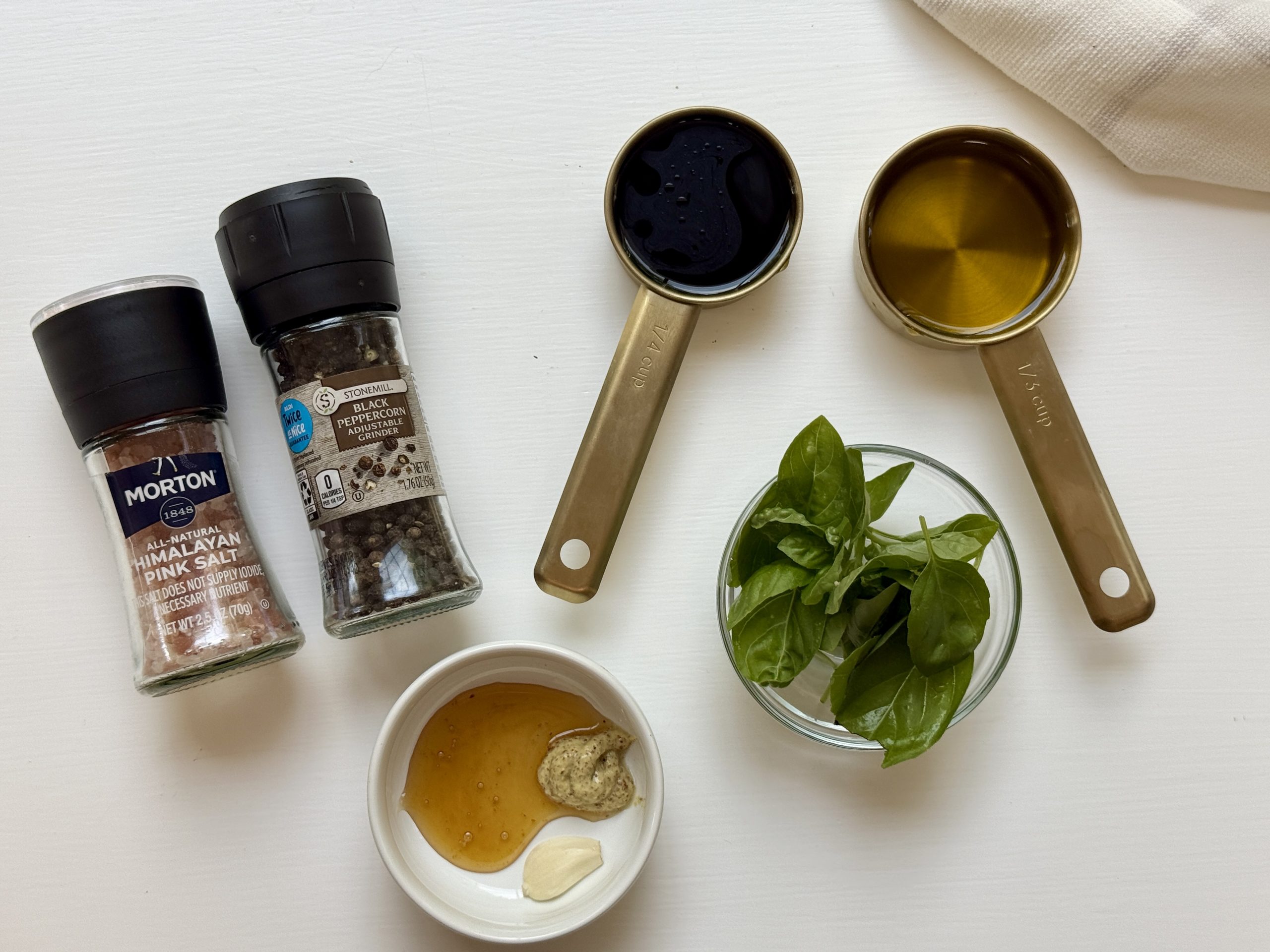 Overhead view of Himalayan pink salt, black pepper, a measuring cup of balsamic vinegar, a measuring cup of olive oil, fresh basil leaves in a small bowl, and a small bowl with honey, Dijon mustard, and garlic.