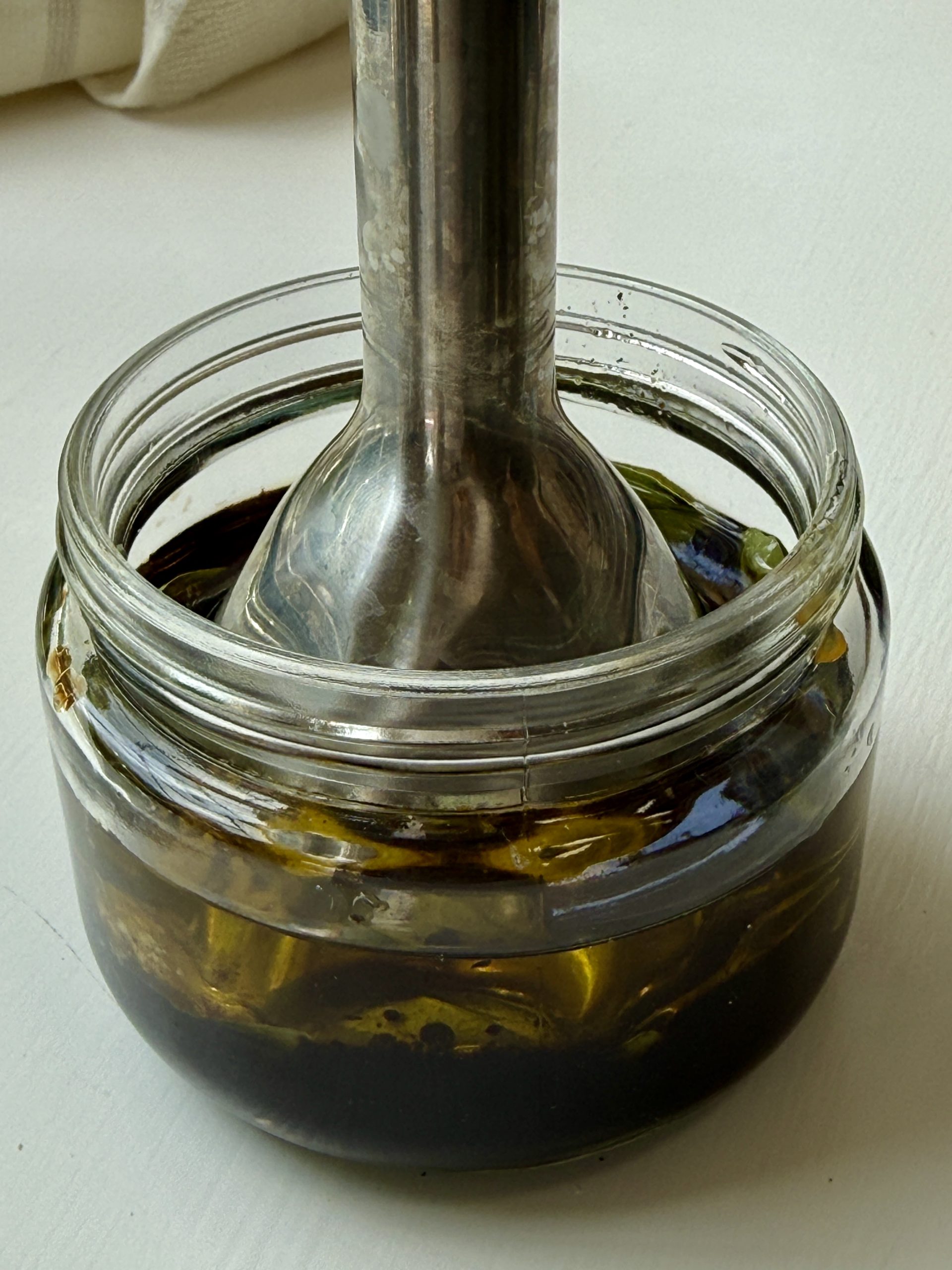 A metal immersion blender is placed inside a glass jar containing layers of dark liquid and olive oil, ready to blend the ingredients.