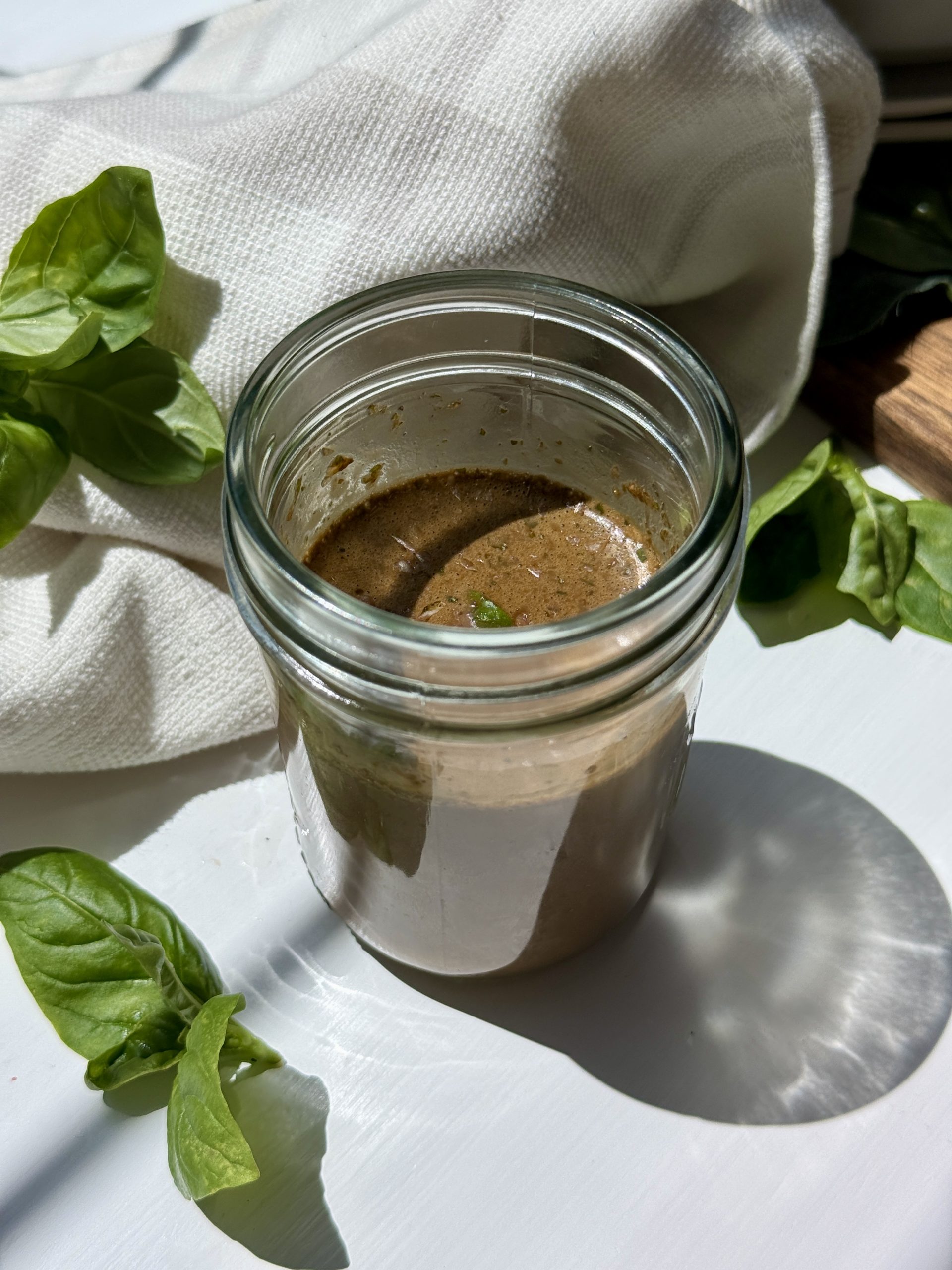 A glass jar filled with brown vinaigrette sits on a white surface, surrounded by fresh basil leaves and a white cloth, with sunlight casting shadows.