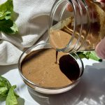 A hand pours brown dressing from a glass jar into a small bowl on a white surface, with fresh basil leaves and a white cloth in the background.