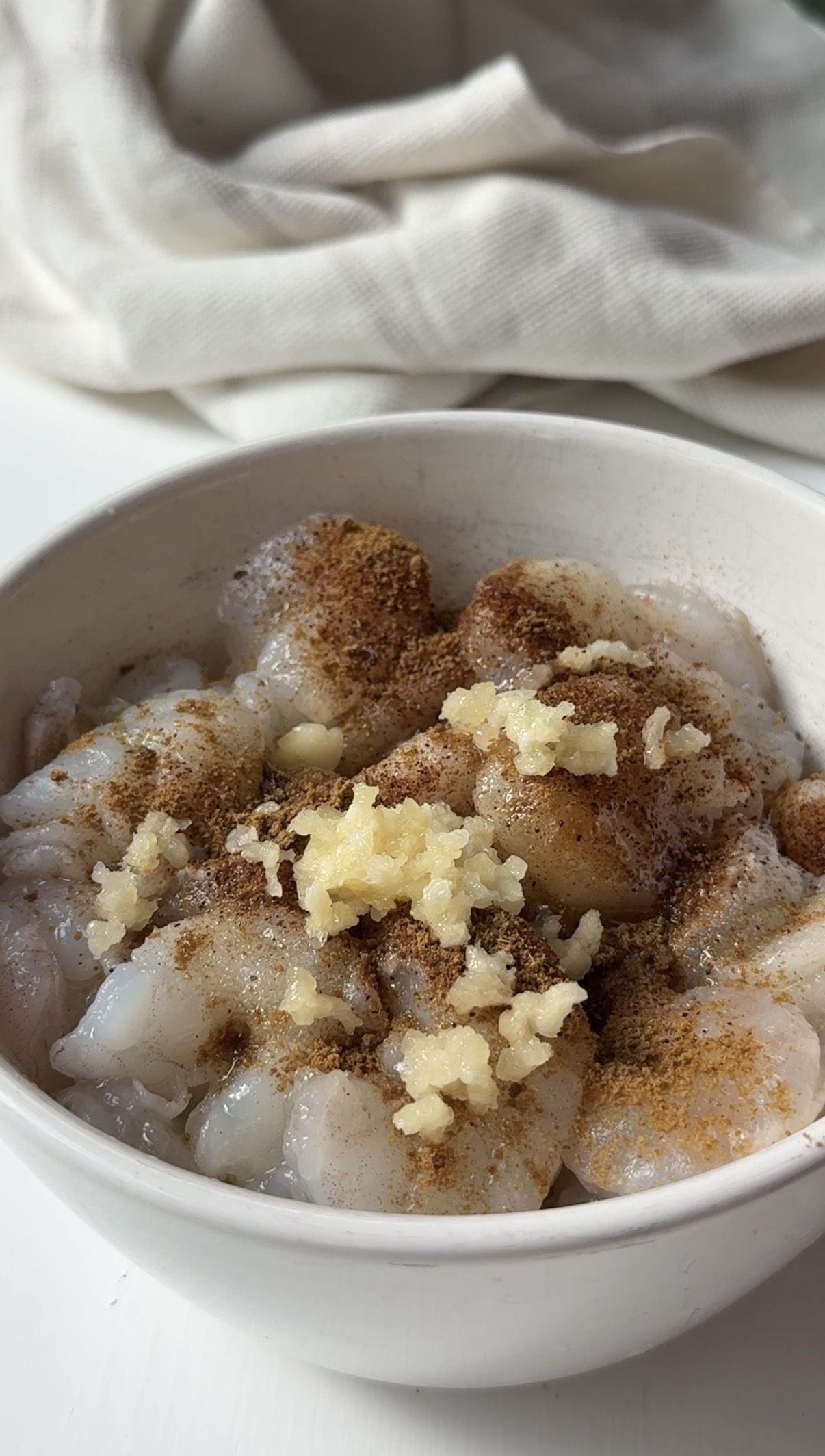 A white bowl filled with raw, deveined shrimp topped with minced garlic and sprinkled spices, sitting on a white surface with a light-colored cloth in the background.