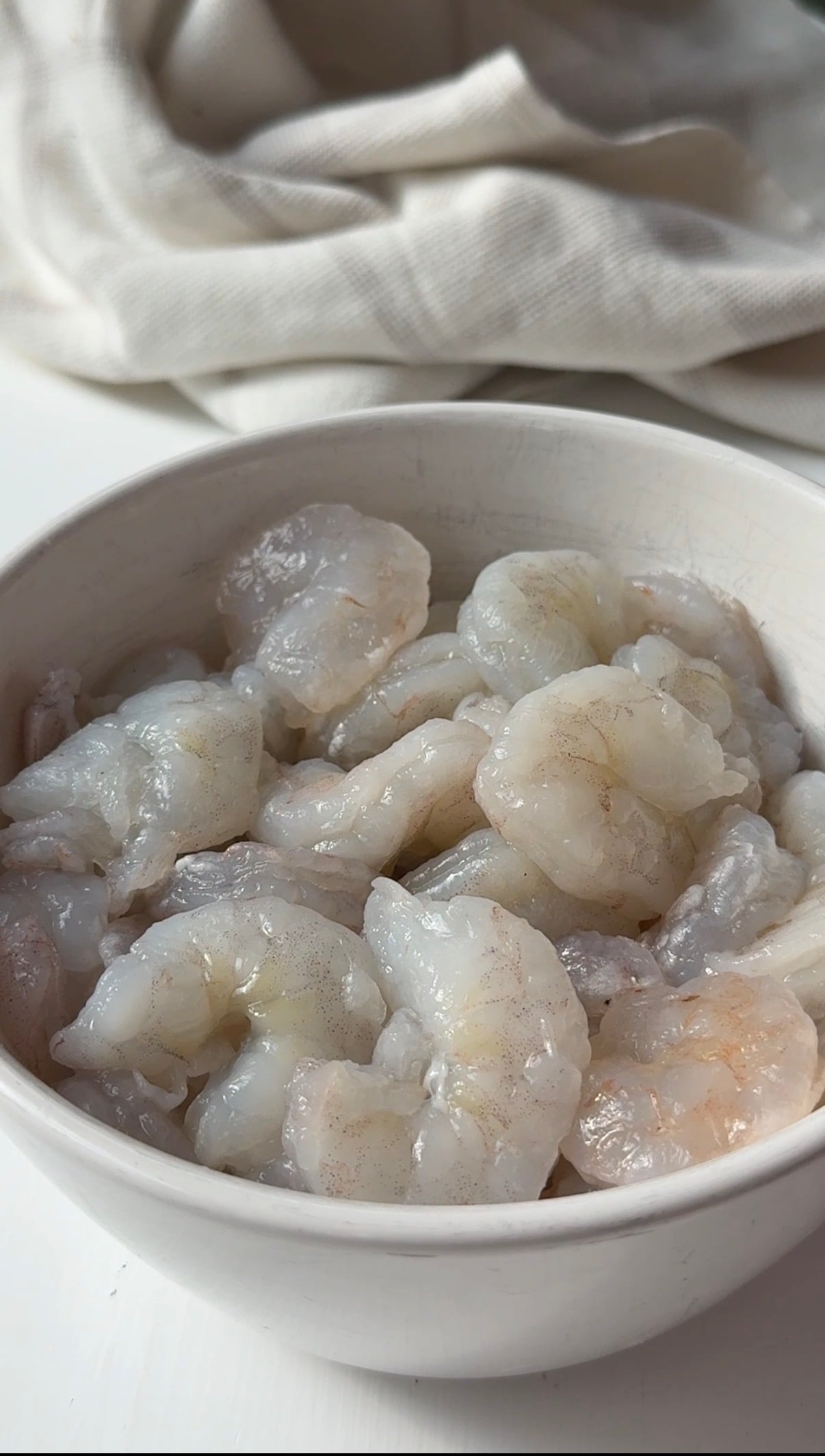 A white bowl filled with raw, peeled shrimp on a white surface.