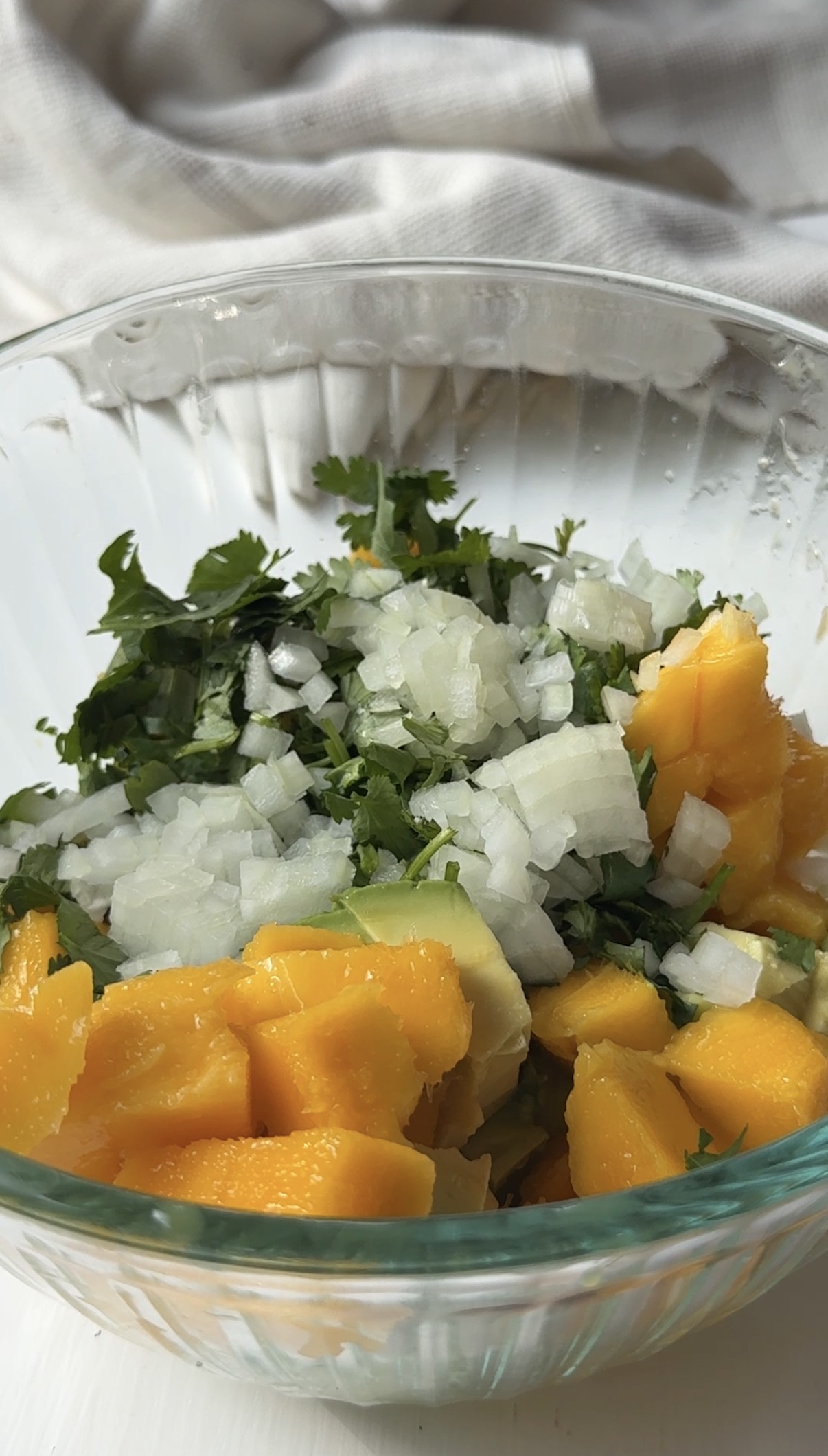 A clear glass bowl filled with chopped mango, avocado, white onion, and fresh cilantro on a white surface, ready to be mixed for a fresh salsa or salad.