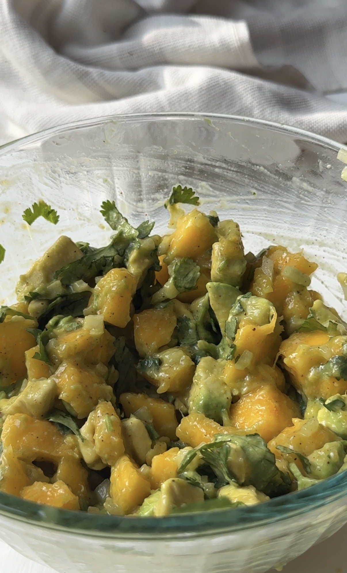 A glass bowl filled with a fresh mango avocado salad, featuring diced mango, avocado, cilantro, and onion. The vibrant yellow and green ingredients are mixed together, with a light dressing visible.