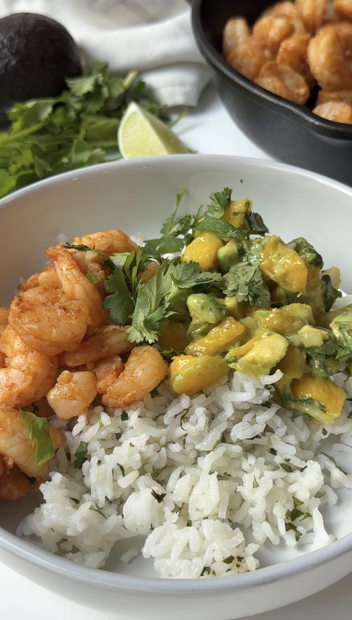 A bowl of white rice topped with seasoned shrimp and a fresh mango avocado salsa, garnished with cilantro. In the background, cilantro, lime, avocado, and a pan of shrimp are visible.