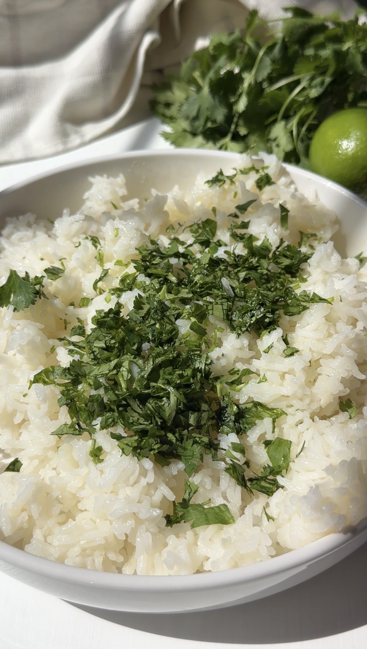 A white bowl filled with cooked white rice, topped with chopped fresh cilantro. In the background, there is a bunch of cilantro and a whole lime on a white surface.