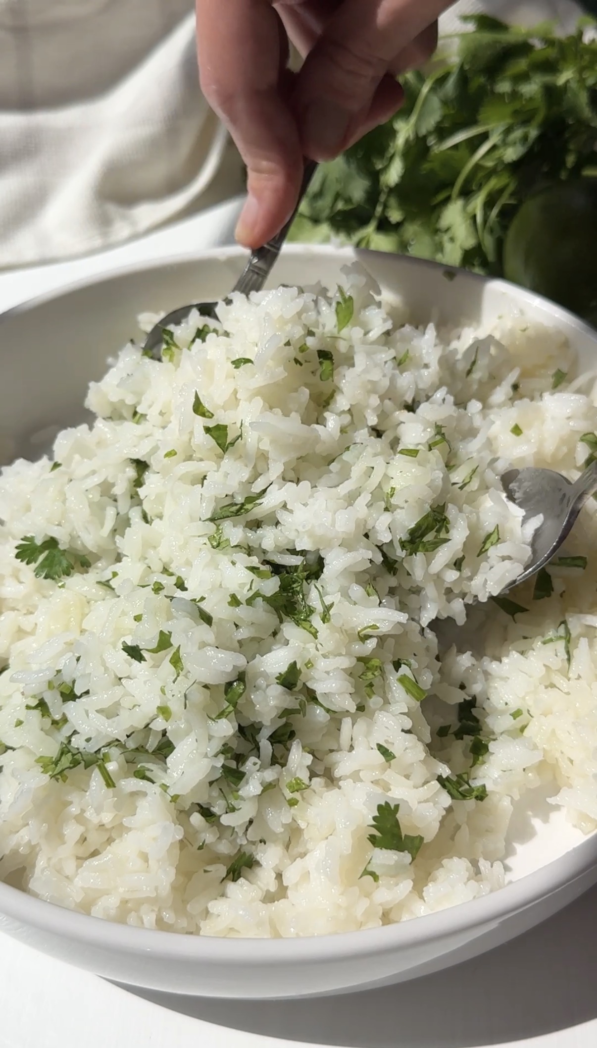A hand uses a spoon to serve fluffy white rice garnished with chopped cilantro from a white bowl; fresh cilantro and a lime are visible in the background.