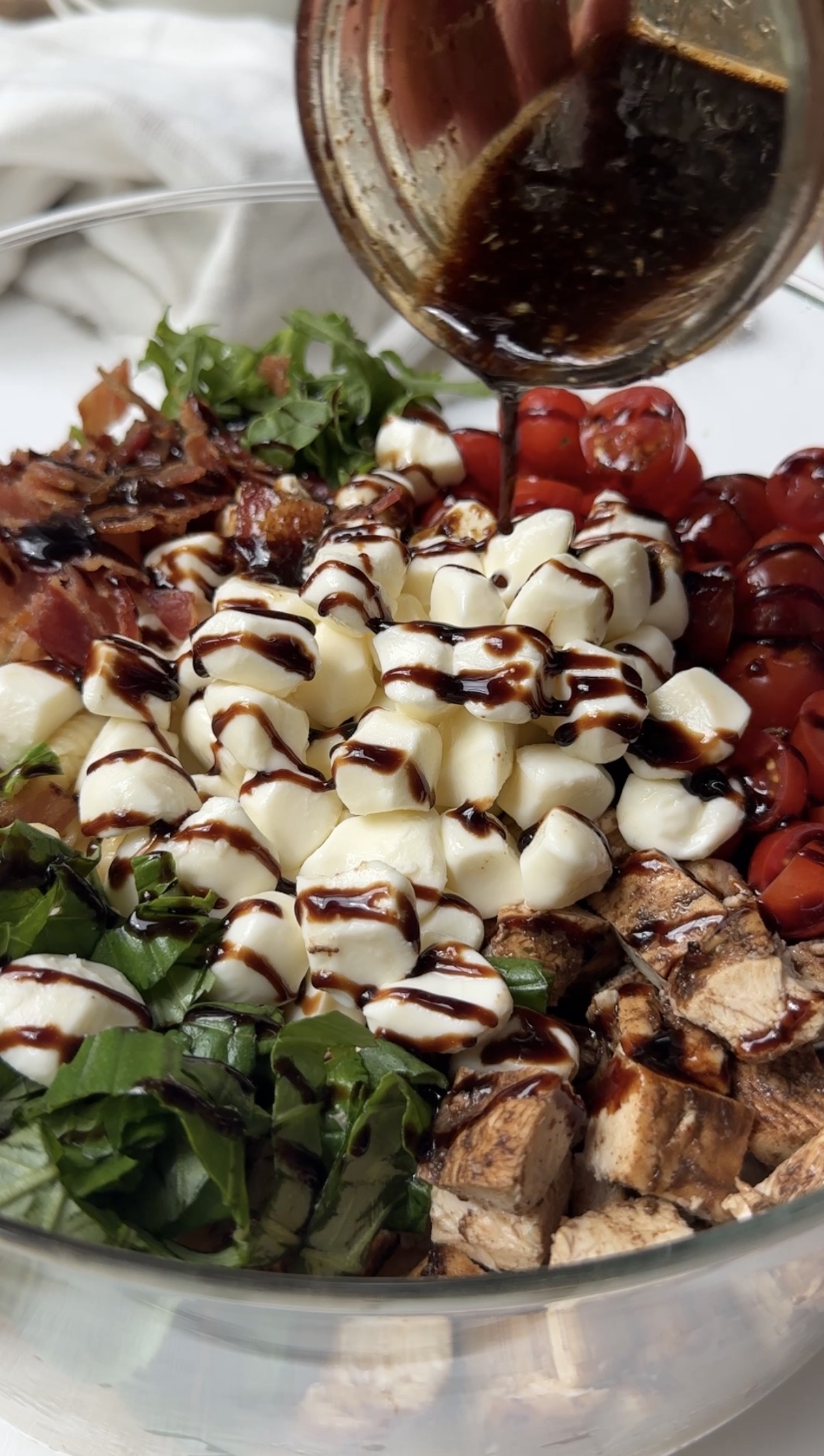 A close-up of a salad with cherry tomatoes, mozzarella balls, grilled chicken, bacon, and greens, drizzled with balsamic glaze being poured from a small dish.