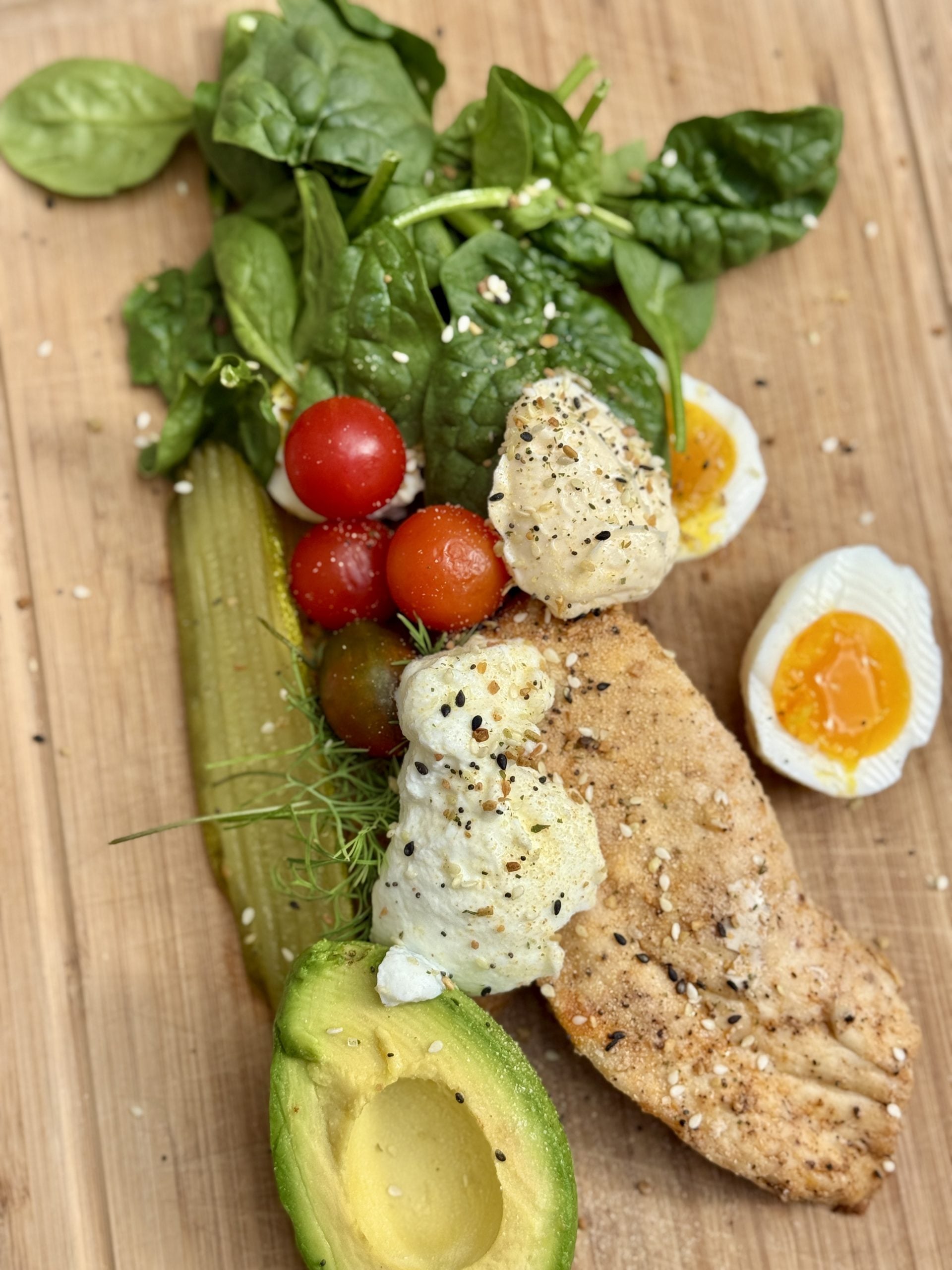 A wooden board topped with a grilled chicken breast, half an avocado, a pickle, cherry tomatoes, fresh spinach leaves, two halves of a soft-boiled egg, and dollops of ricotta cheese, sprinkled with seasoning.