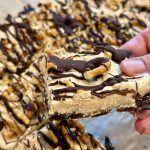 A hand holds up a square dessert bar topped with chocolate drizzle and pretzels, with more bars arranged on parchment paper in the background.