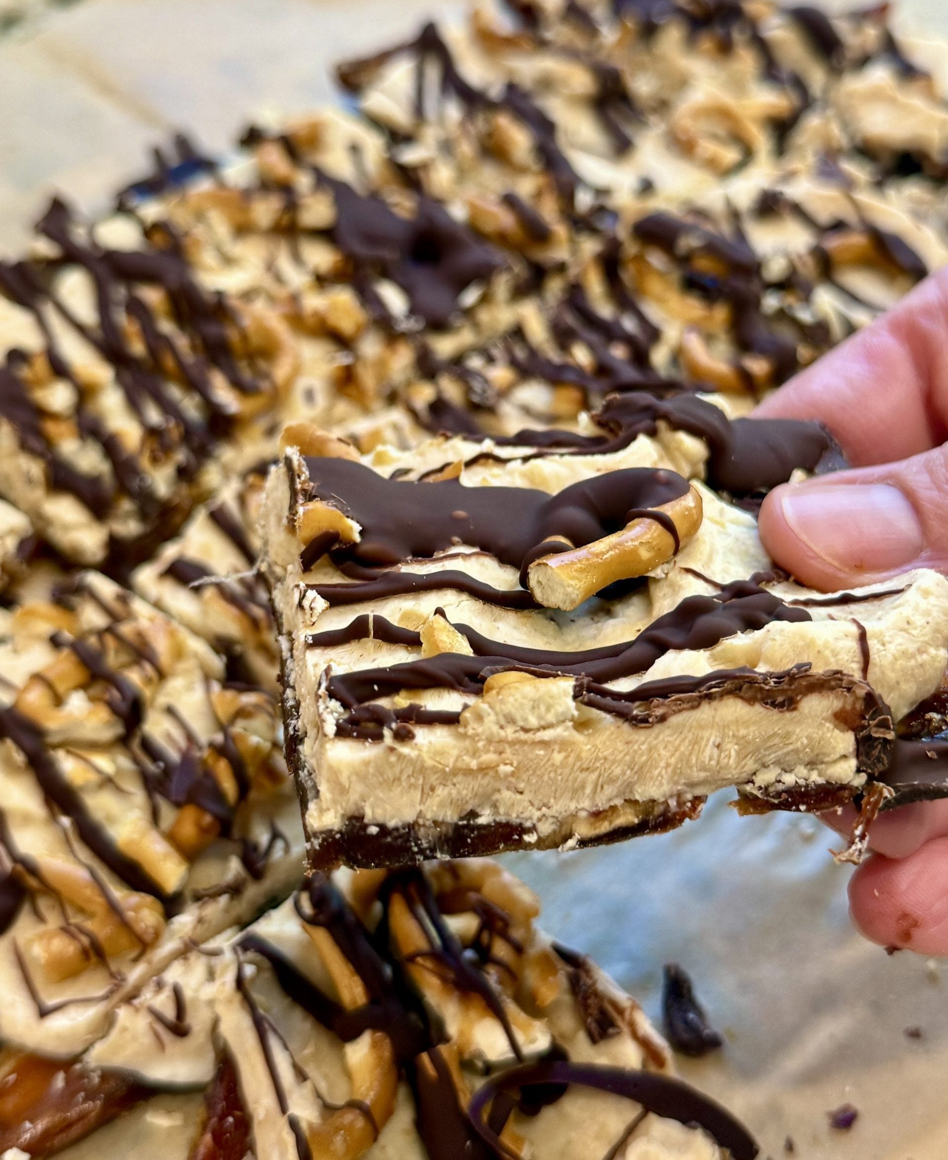 A hand holds up a square dessert bar topped with chocolate drizzle and pretzels, with more bars arranged on parchment paper in the background.