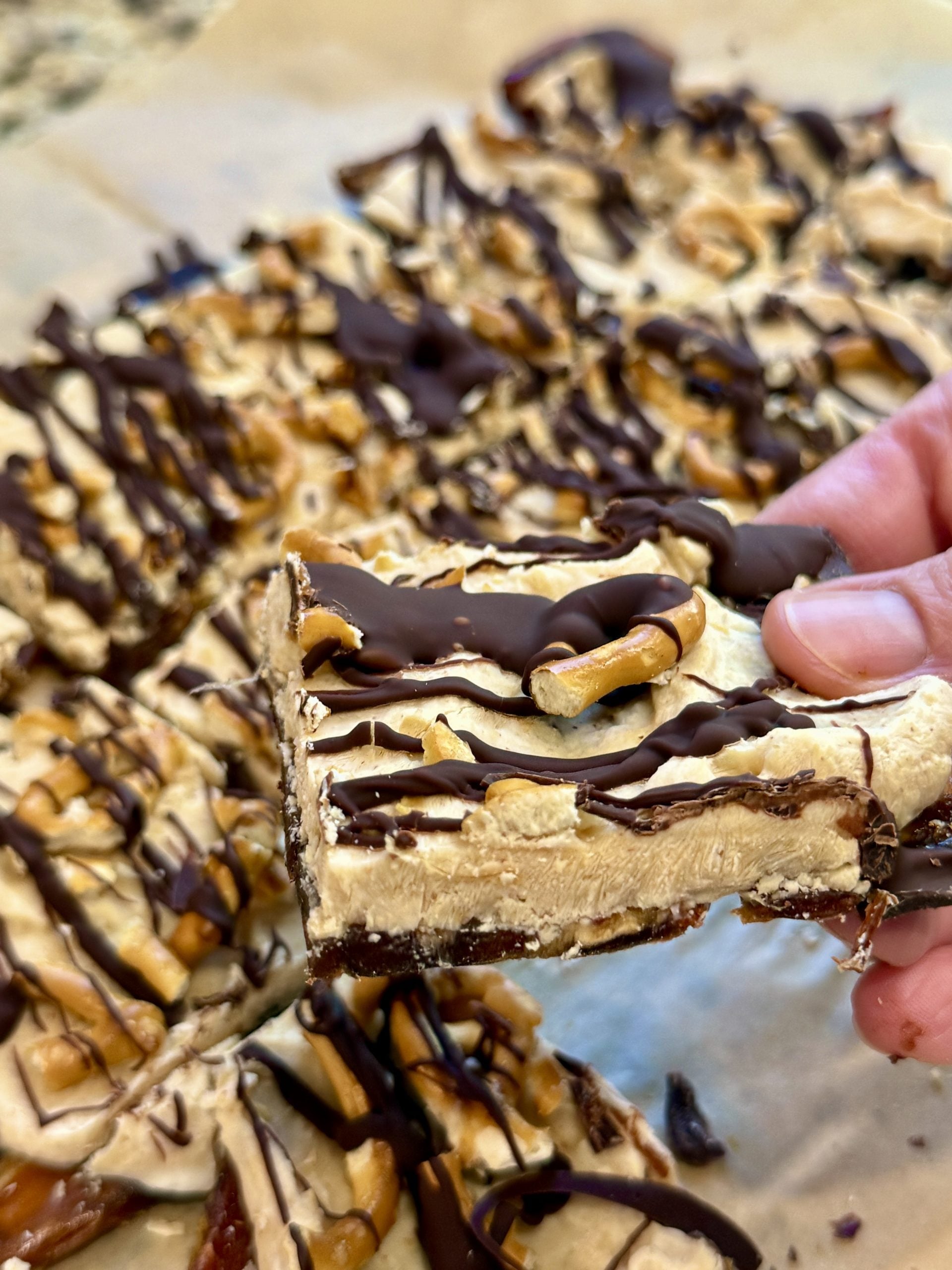 A hand holds up a square dessert bar topped with chocolate drizzle and pretzels, with more bars arranged on parchment paper in the background.
