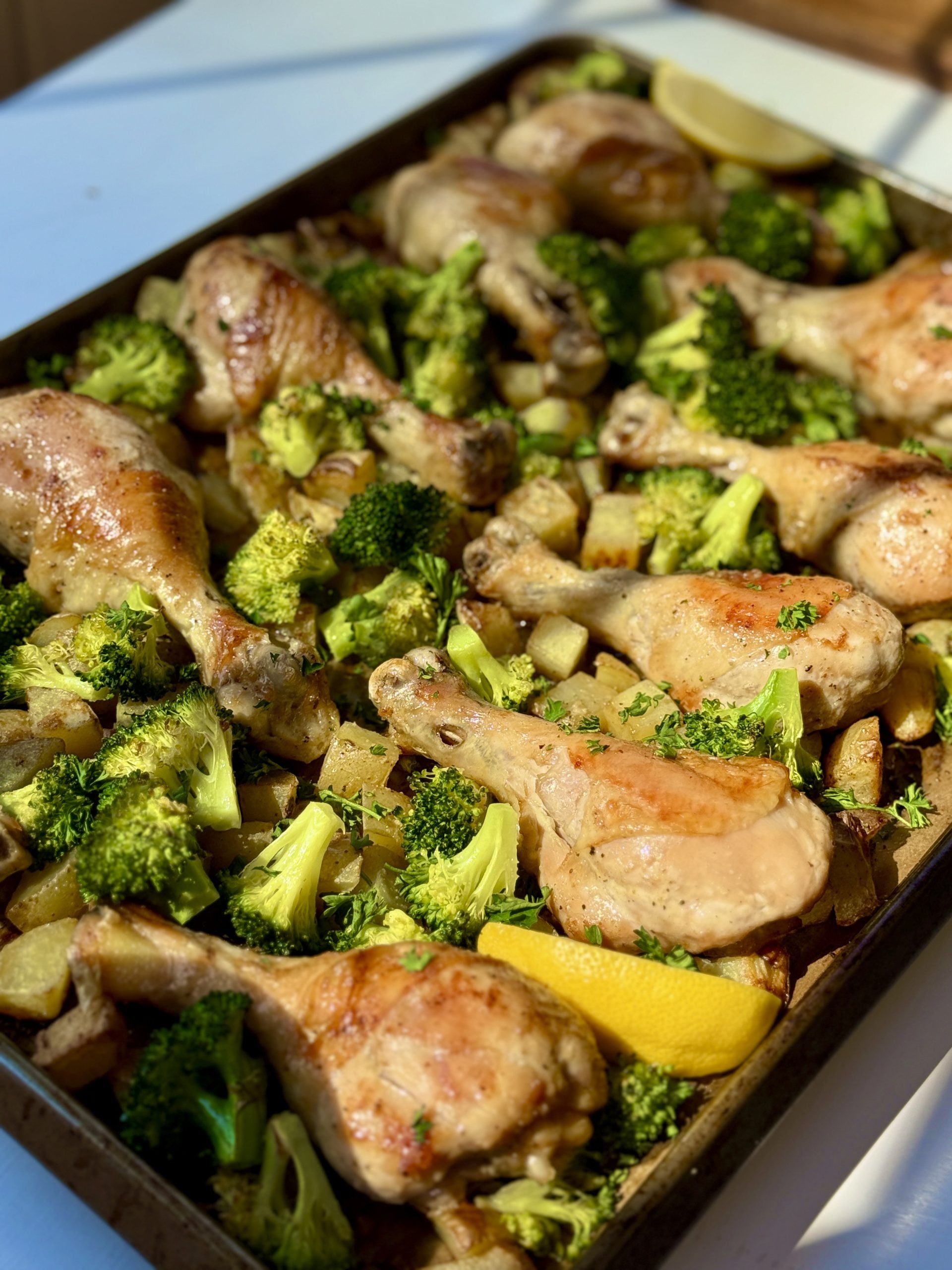 A baking tray filled with roasted chicken drumsticks, broccoli florets, and potato pieces, garnished with lemon wedges under natural sunlight.