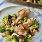 A plate with two roasted chicken drumsticks, surrounded by roasted broccoli and diced potatoes, with a baking tray of more chicken and veggies in the background.